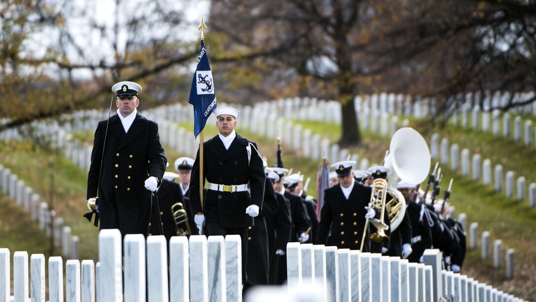 Sailors walk in formation through rows of white headstones.