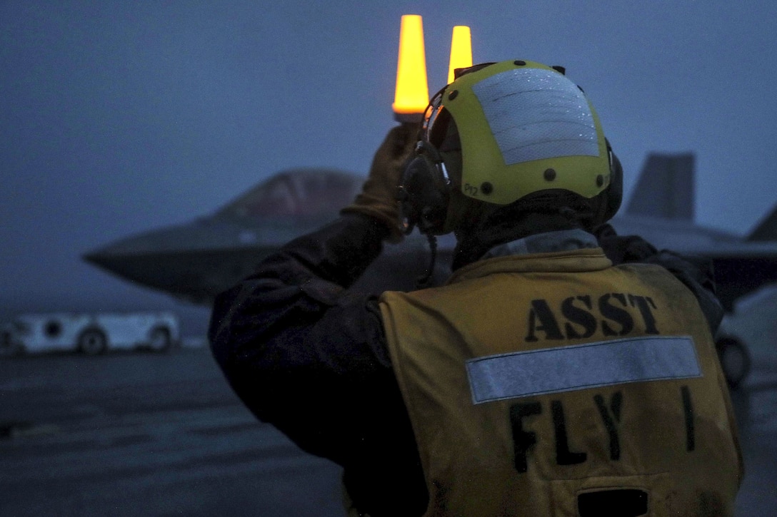 A sailor holds two yellow lights above his face on a ship's flight deck, with an jet in the background.