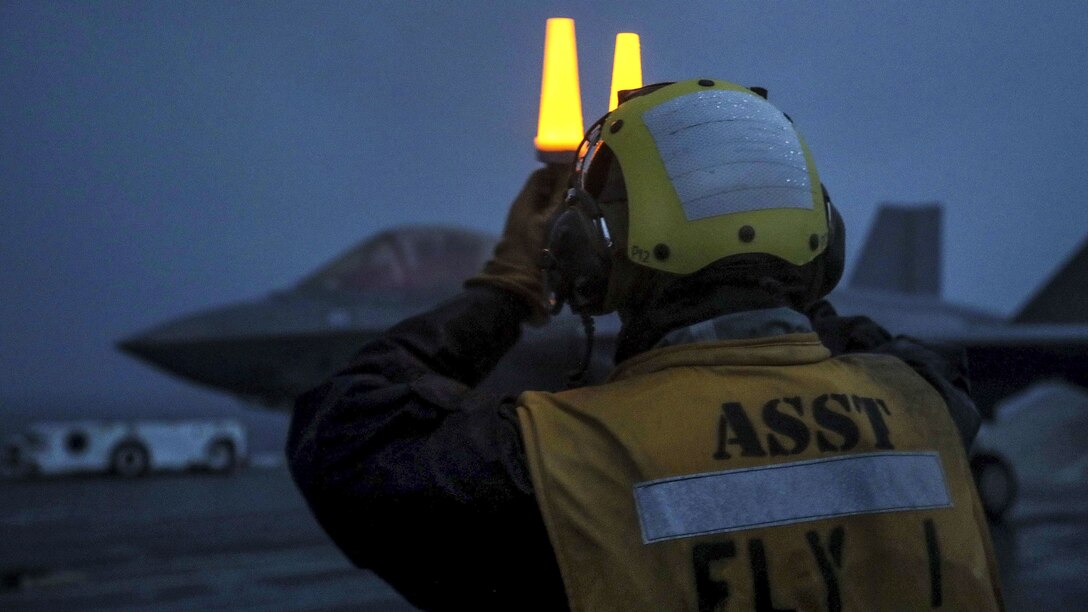 A sailor holds two yellow lights above his face on a ship's flight deck, with an jet in the background.