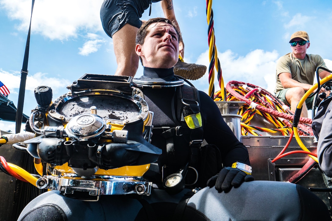 A sailor sits while someone else helps him put on gear.