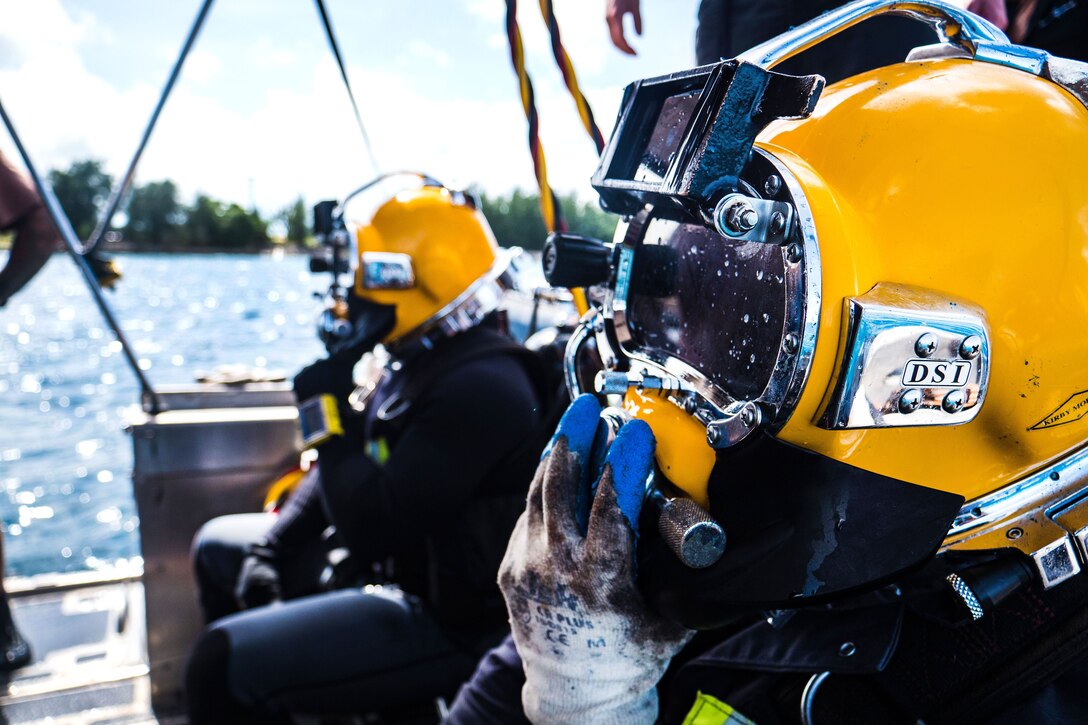 Sailors in diving gear hold their hands up to their helmets.