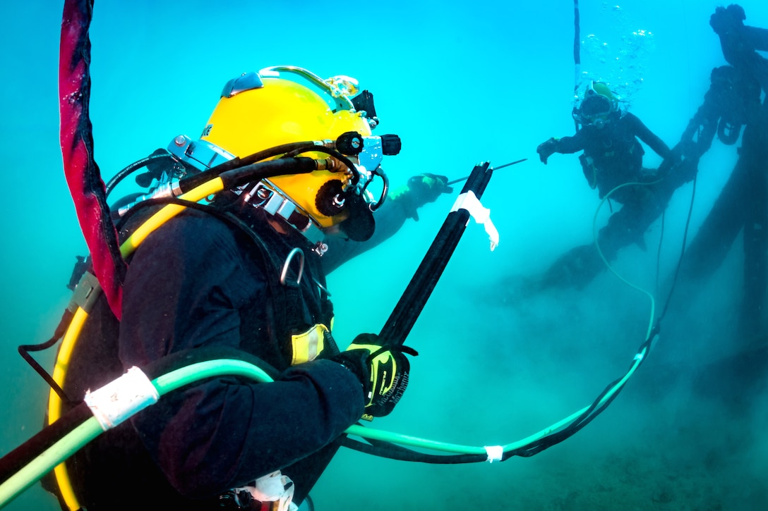 A diver hands equipment to another diver while underwater.