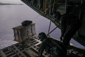 Two service members push a package off the back of an aircraft in flight.
