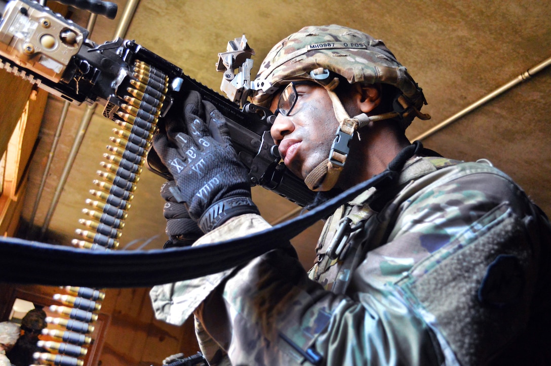 A soldier holds a firearm while looking out a window.
