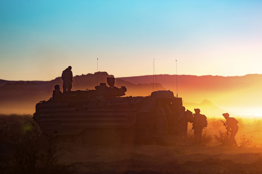 Marines, shown in silhouette against blue and orange sky, board an amphibious vehicle on sandy terrain.