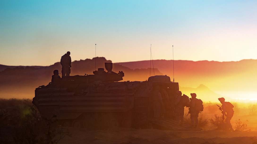 Marines, shown in silhouette against blue and orange sky, board an amphibious vehicle on sandy terrain.