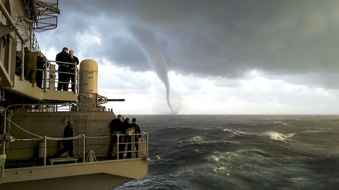 Sailors aboard a ship look out as a funnel shape cloud forms over the sea.
