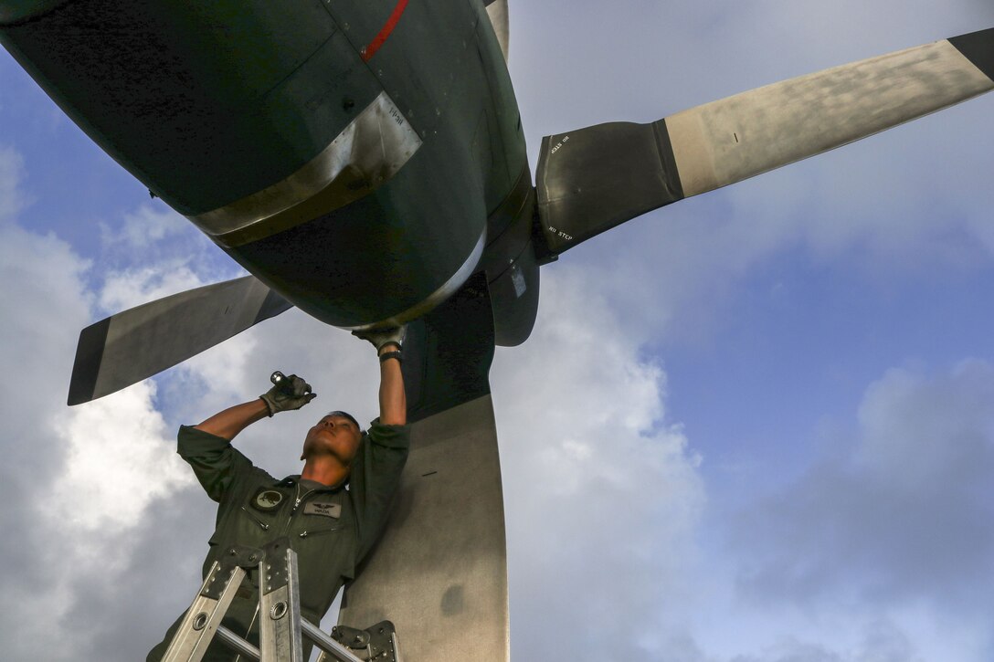 An airman on a ladder reaches and looks up at an aircraft propeller he's inspecting.