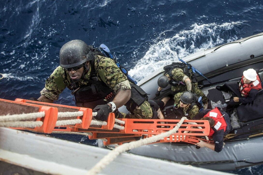 Sailors, viewed from above, climb an orange-stepped rope ladder from an inflatable boat up the side of a ship.