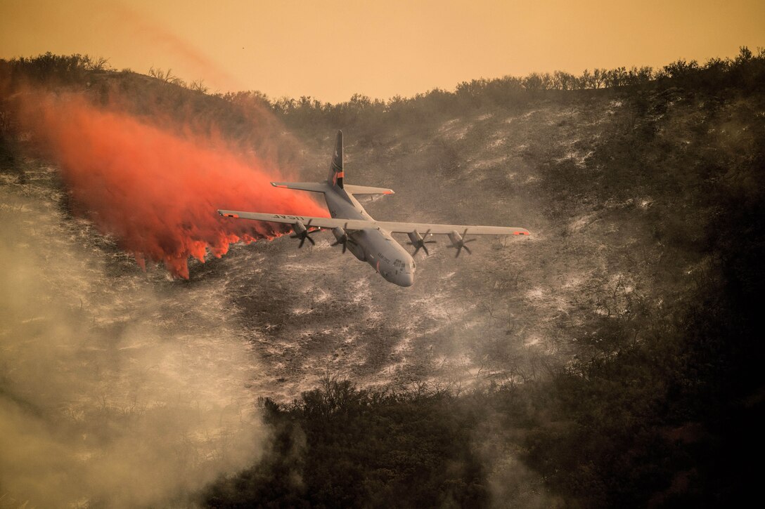 Orange smoke streams from an aircrfat flying over hilly terrain.