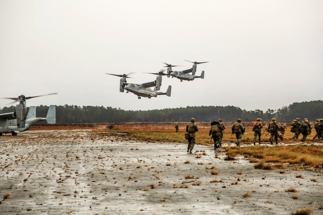 Marines run on the ground with aircraft overhead.