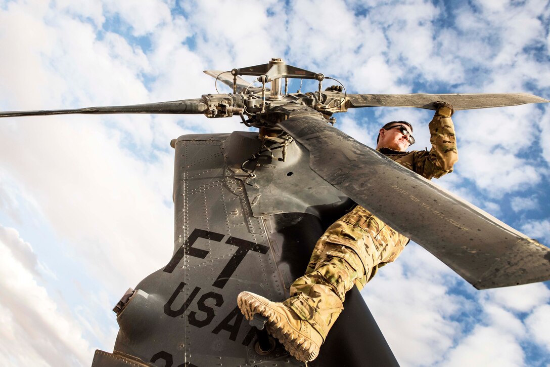 An airman sits on the tail of a helicopter looking at the rotor blades.