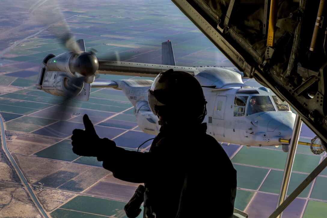 A Marine, shown in profile and silhouette, gives a thumbs up aboard an aircraft in flight near a helicopter.