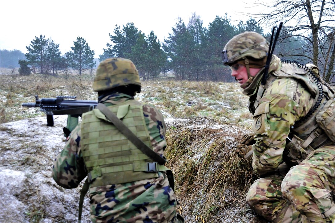 A U.S. soldier sits next to a Polish soldier firing a weapon.