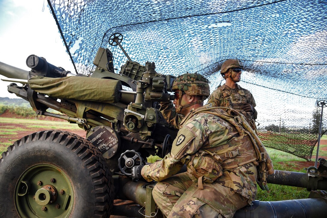 Two soldiers sit and stand next to a howitzer gun.