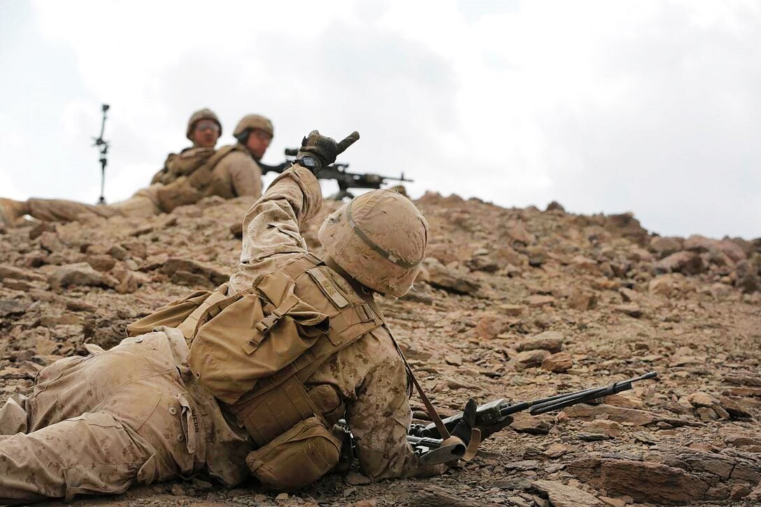 A Marine lays on the ground and makes a hand signal.