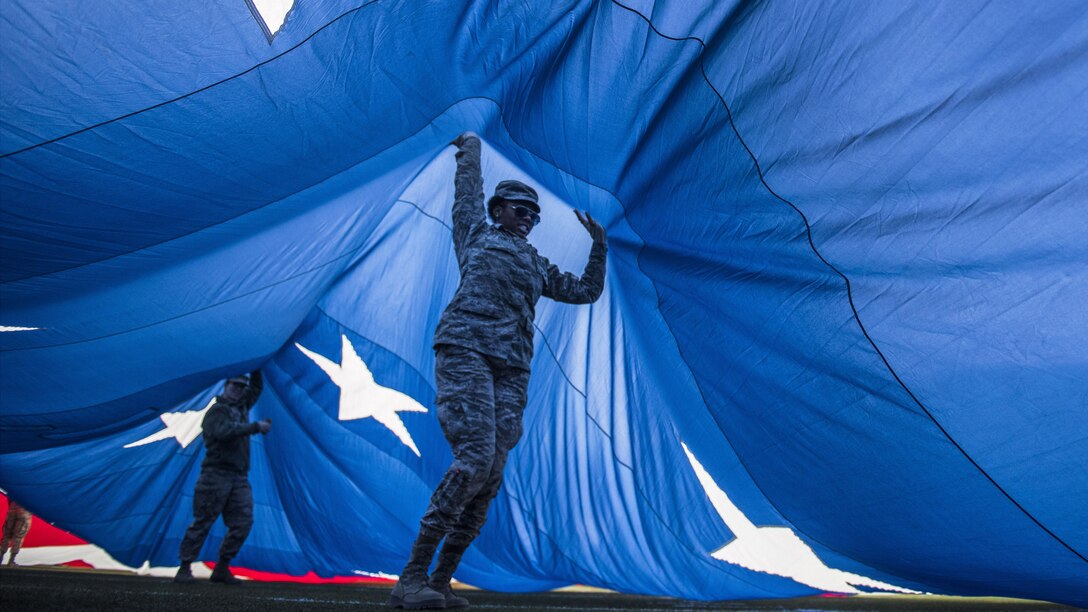Two airmen hold up the blue section of a giant U.S. flag while standing underneath it.