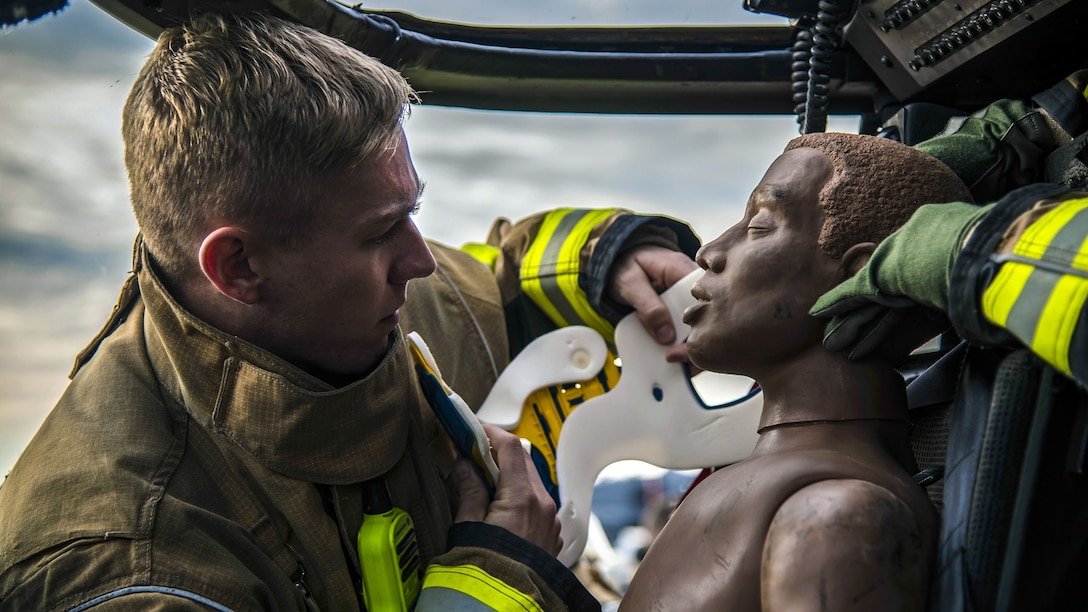 An airman faces a mannequin in a vehicle and puts a neck brace on it.