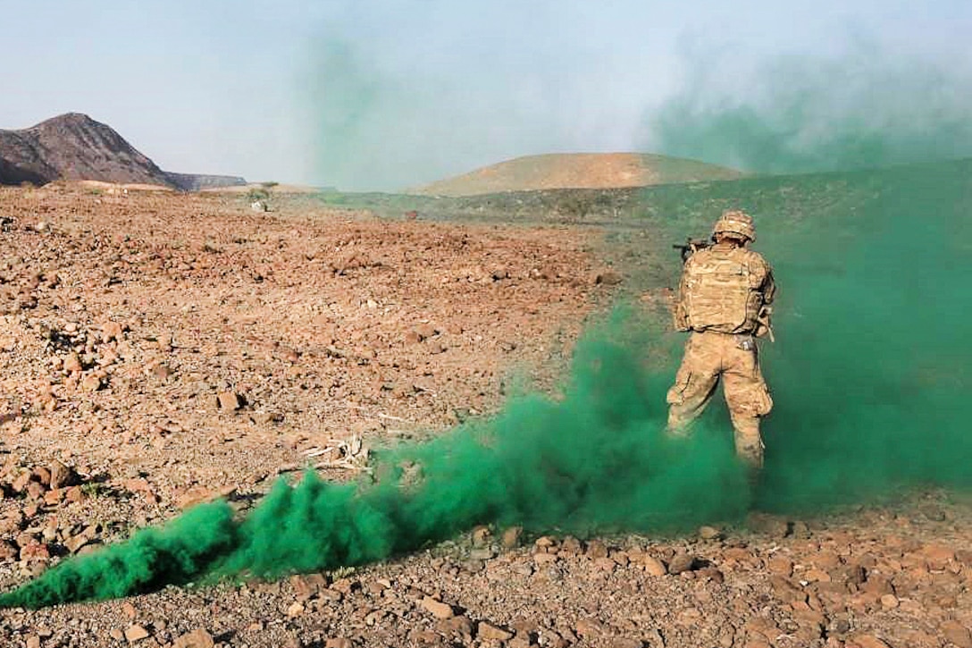 A soldier standing in wafting green smoke stands and aims a weapon in desert terrain.
