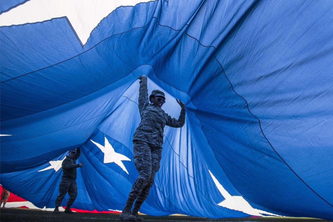 Two airmen hold up the blue section of a giant U.S. flag while standing underneath it.