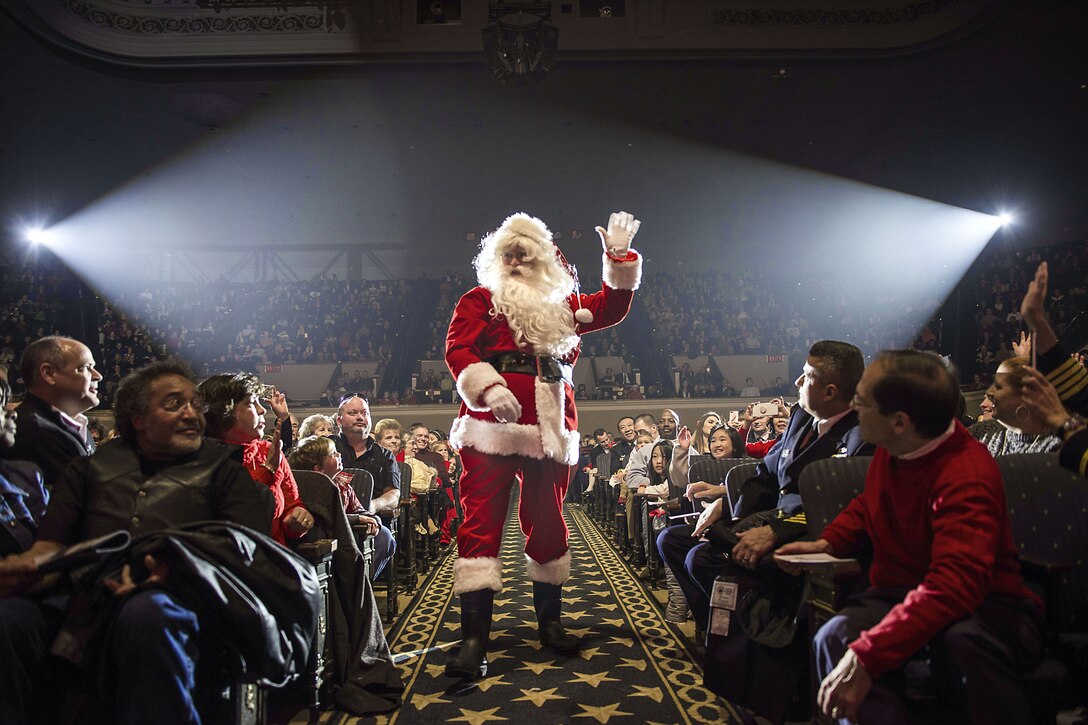 Santa Claus waves to audience members while walking down the center aisle of a concert hall.