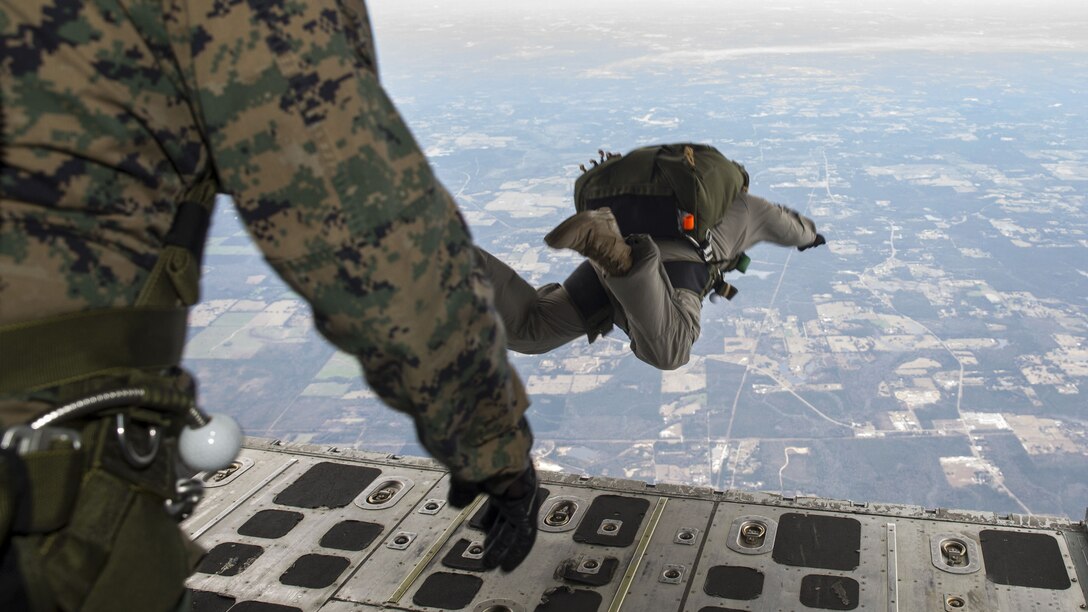 A Marine with an unopened parachute hovers in the air after jumping from an opened aircraft, where another Marine is standing.