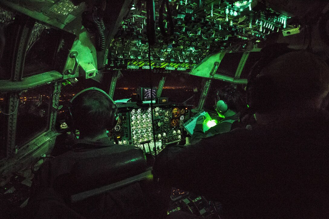 Air Force pilots sit in a cockpit under green lights as they fly an aircraft.