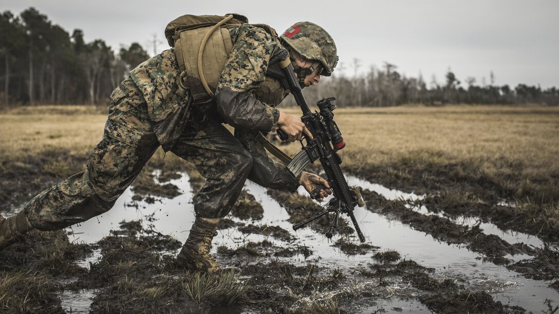 A Marine crouches over as he rushes with his weapon through muddy water.