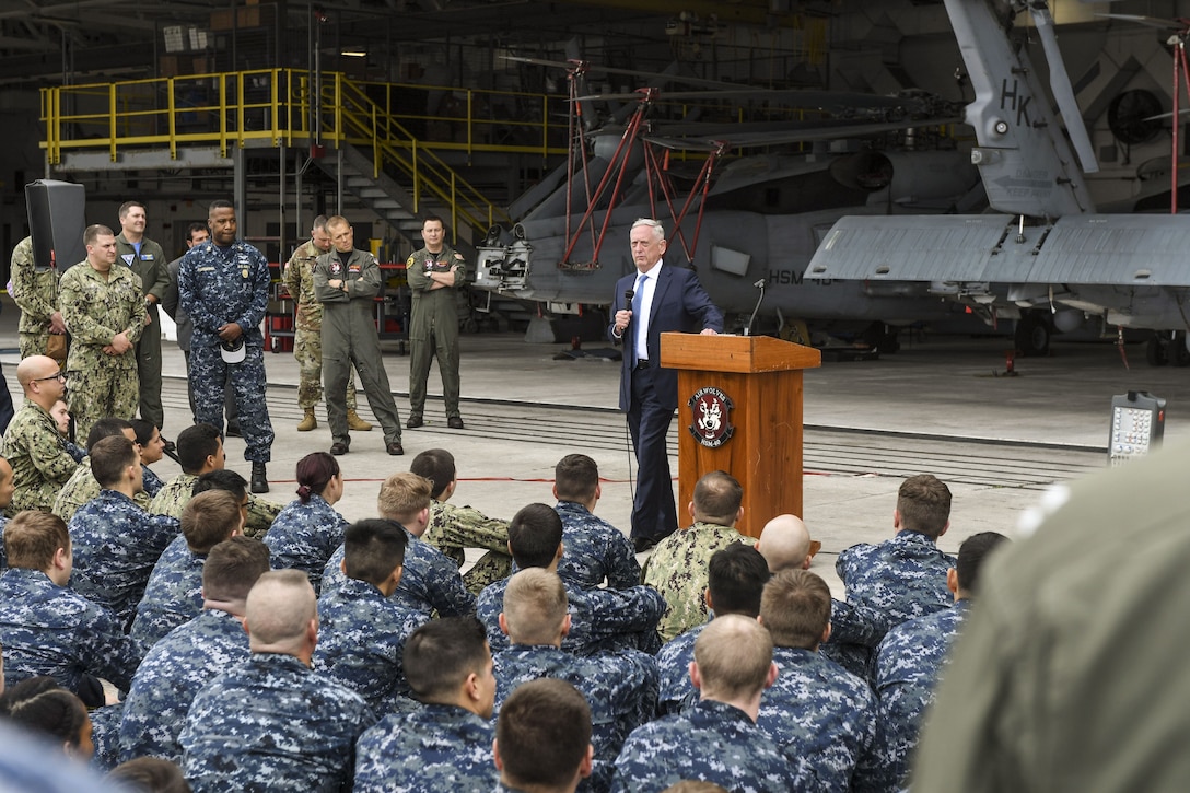 Defense Secretary James N. Mattis stands beside a lectern in a hangar and addresses sailors.