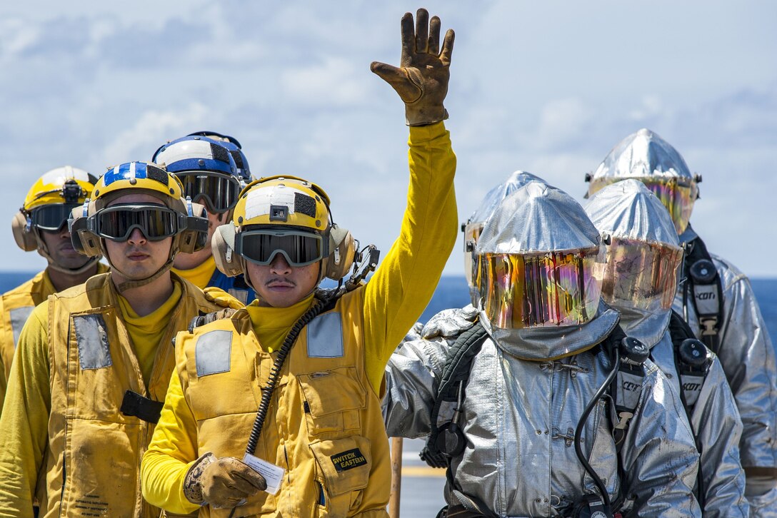 Sailors in yellow and silver suits stand as one waves during firefighting drills.