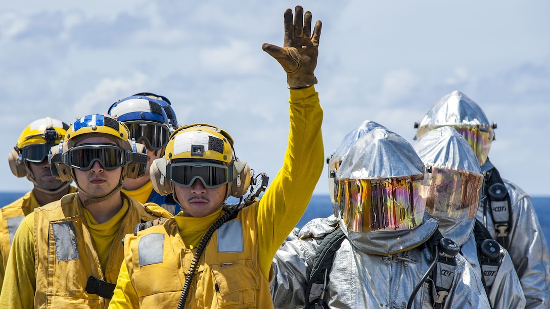 Sailors in yellow and silver suits stand as one waves during firefighting drills.