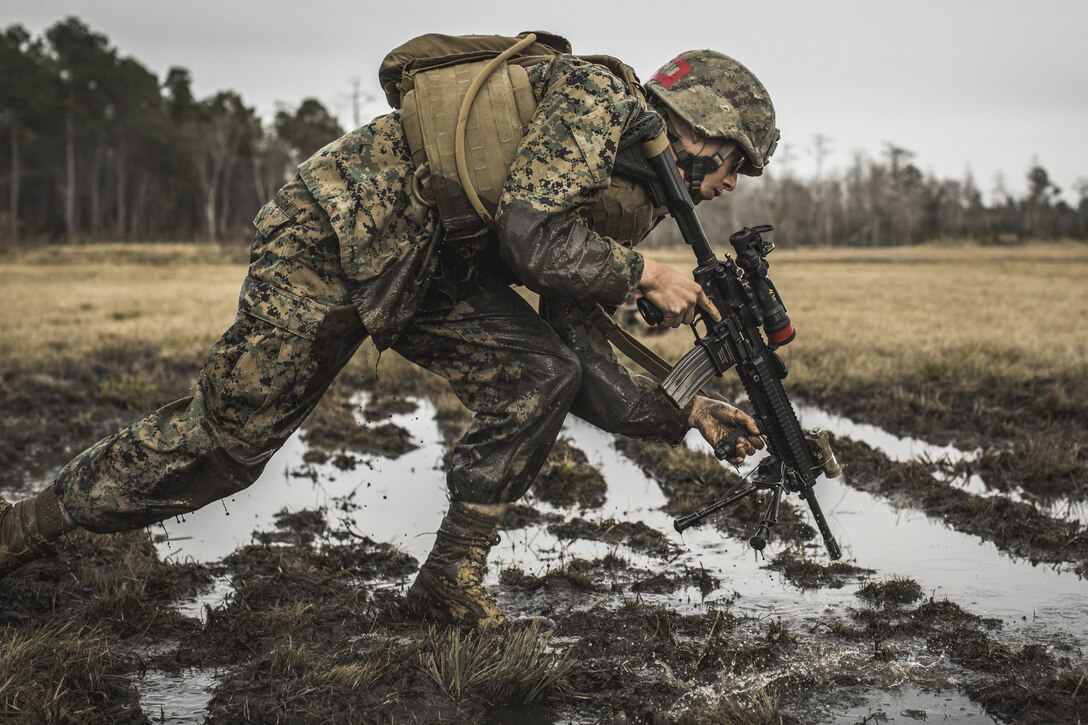 A Marine crouches over as he rushes with his weapon through muddy water.