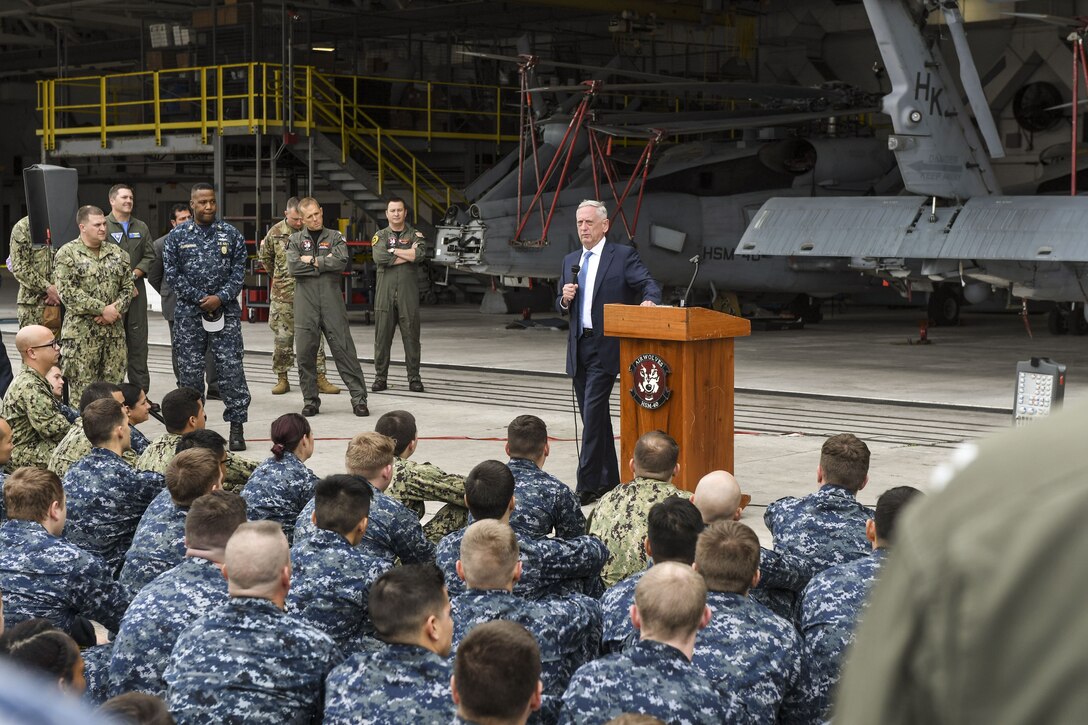 Defense Secretary James N. Mattis stands beside a lectern in a hangar and addresses sailors.