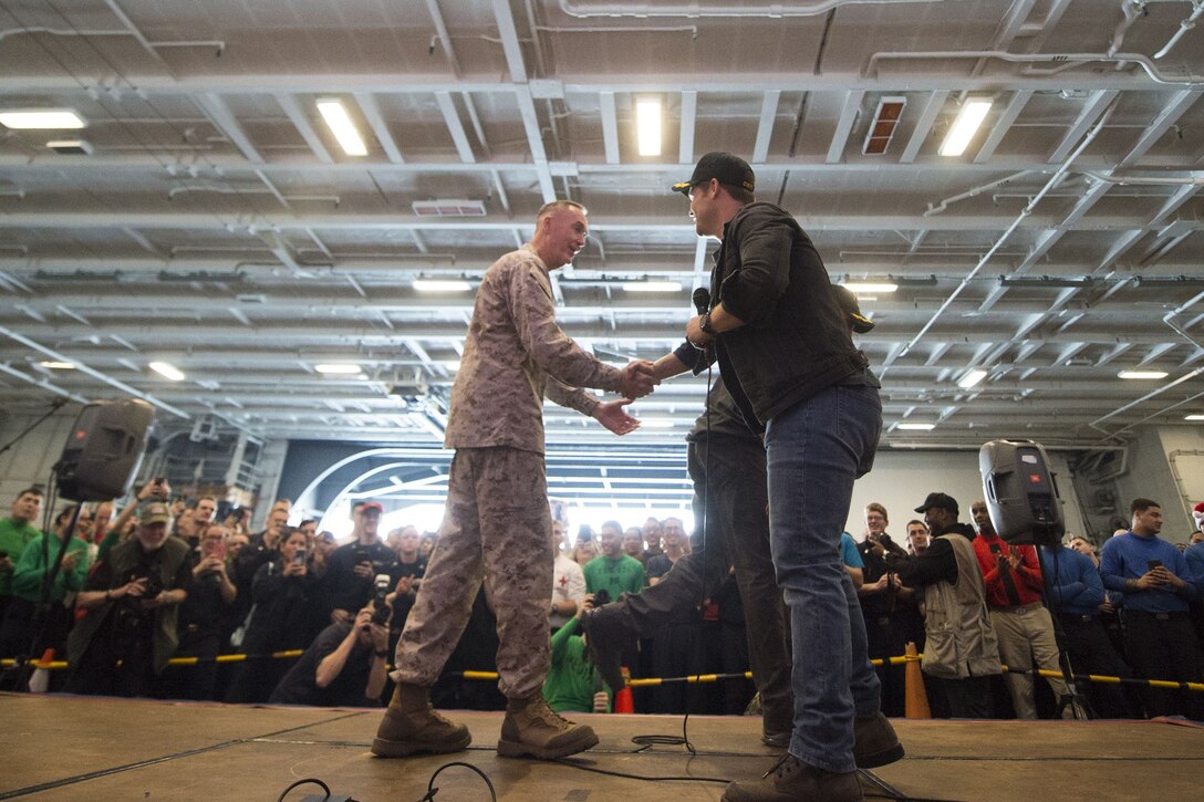 Marine Corps Gen. Joe Dunford shakes hands with Adam Devine on a stage as an audience of sailors watches.