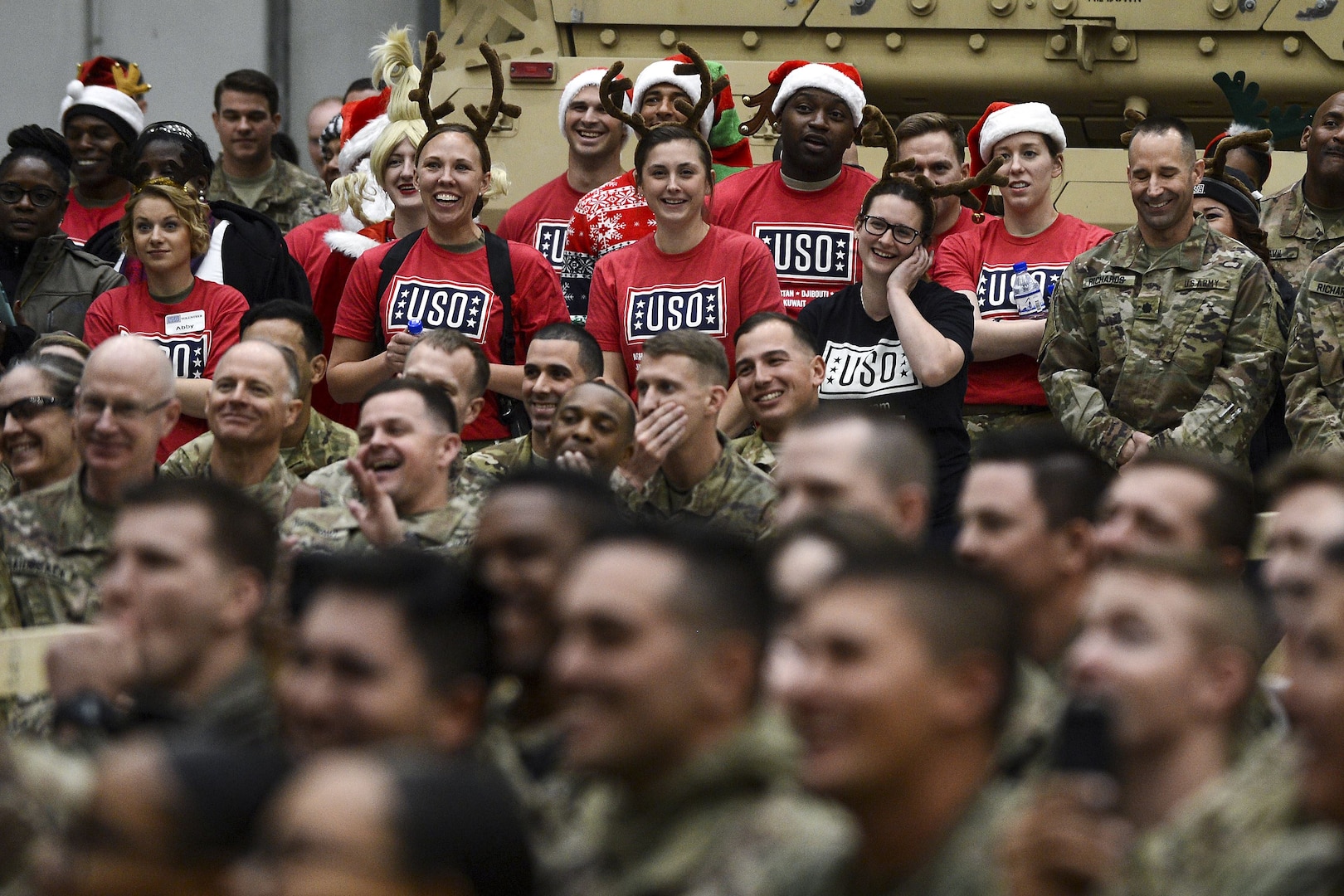 An audience of service members, some in USO shirts and holiday garb, laugh.