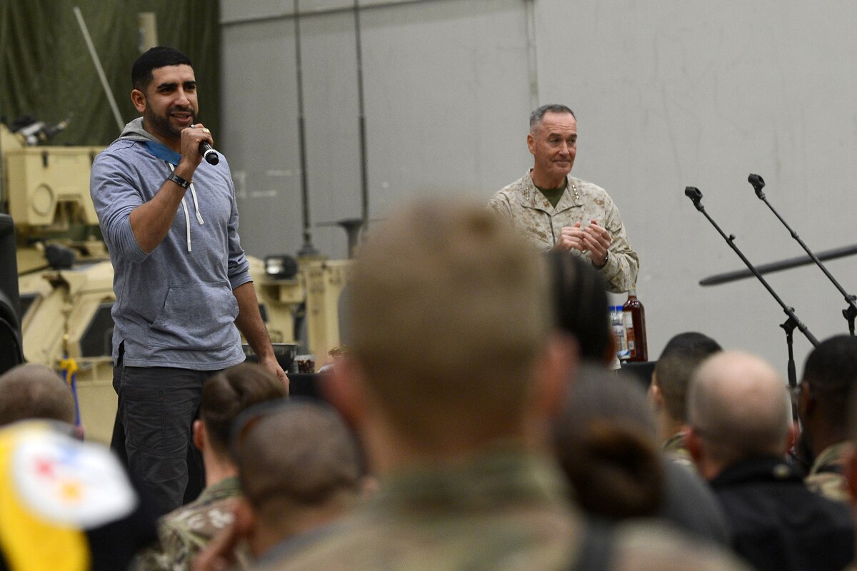 A man speaks into a microphone to a crowd of troops, while Marine Corps Gen. Joe Dunford joins him on stage.