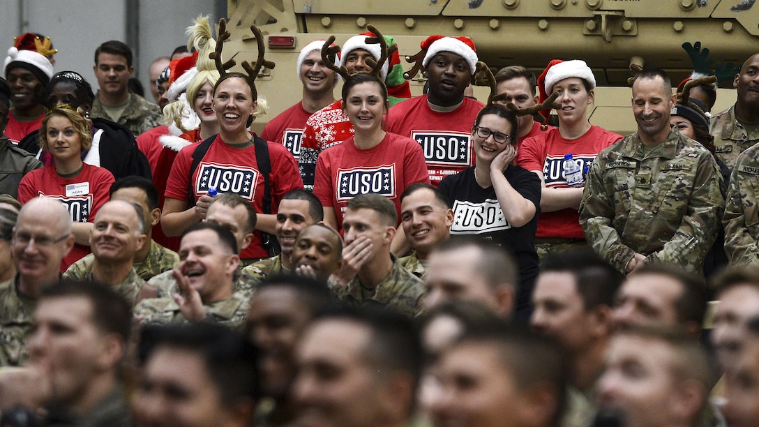 An audience of service members, some in USO shirts and holiday garb, laugh.