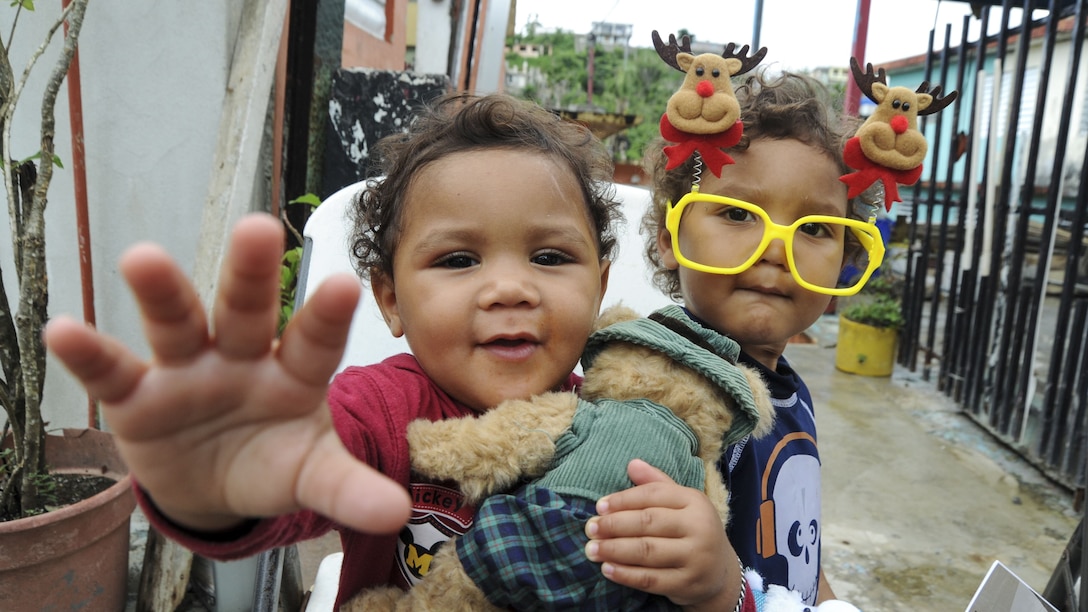 A child holding a stuffed animal reaches toward the camera, while another one wearing Christmas novelty glasses looks on.