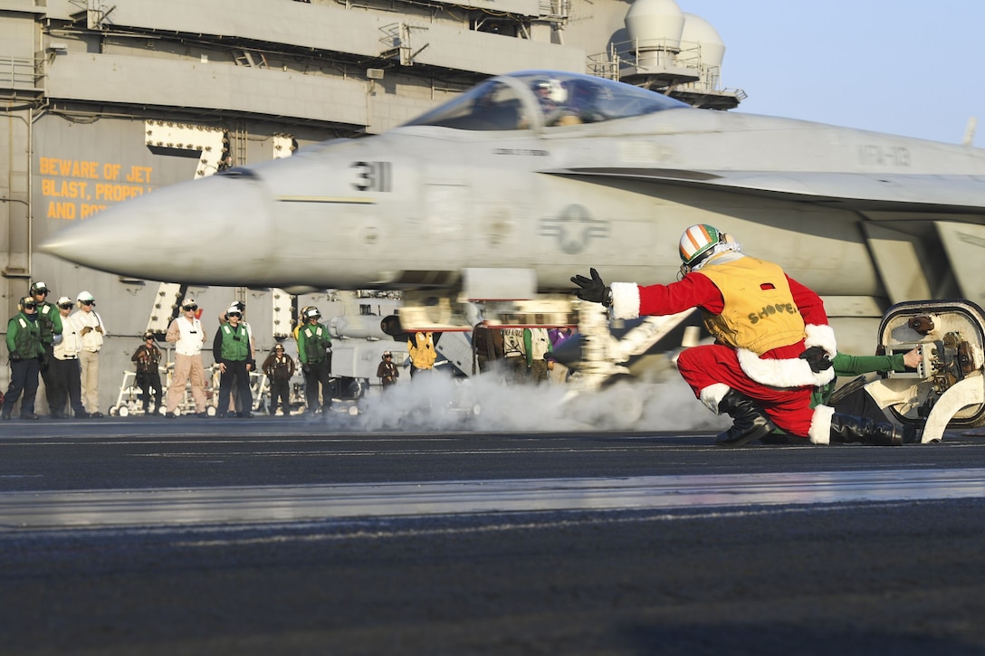 A sailor dressed as Santa Claus kneels and extends an arm as an aircraft takes off from a ship's flight deck.