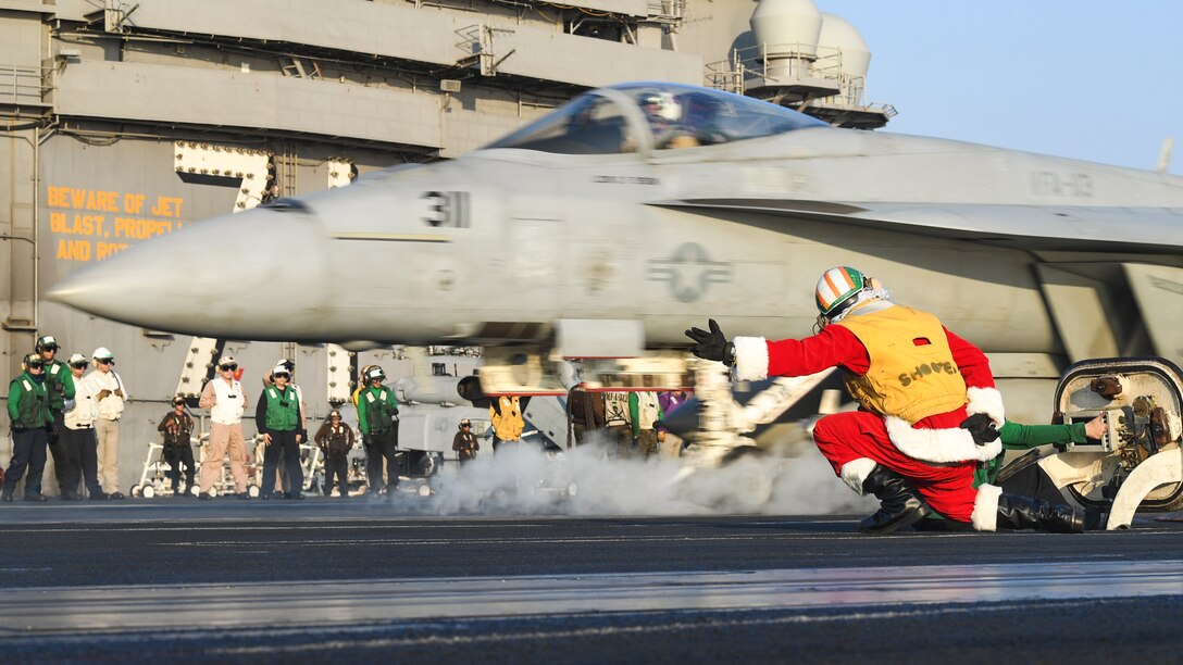 A sailor dressed as Santa Claus kneels and extends an arm as an aircraft takes off from a ship's flight deck.
