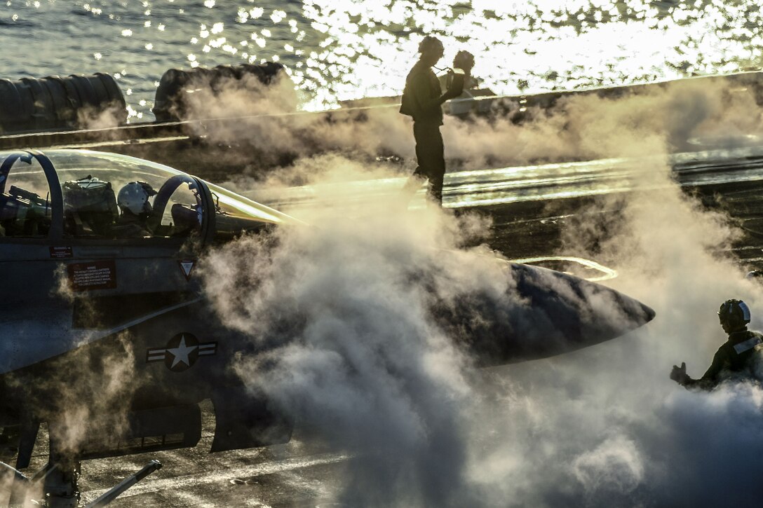 An aircraft and airmen sit on the deck of a ship surrounded by steam.