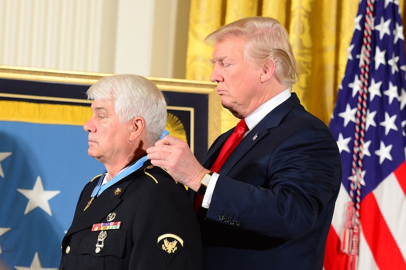 A man in a suit places a medal around the neck of another man in a military dress uniform.