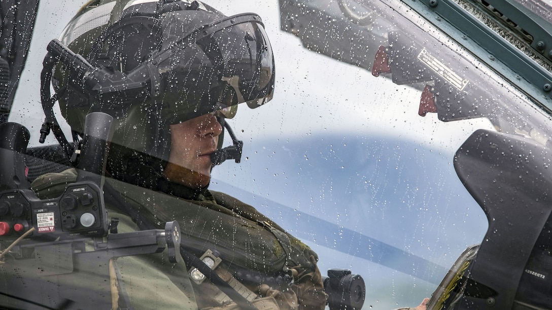 A pilot sits in a helicopter cockpit with water droplets on the windshield.