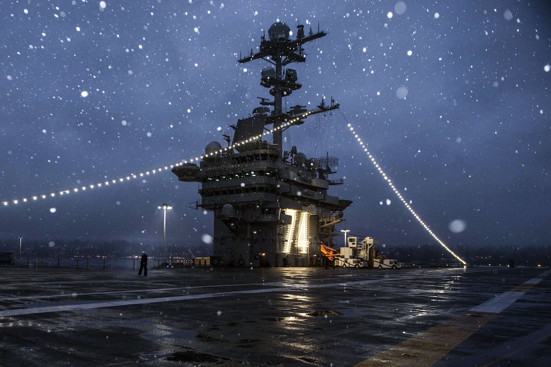 Snowflakes, prominent against a dark blue sky, fall on a ship's flight deck.