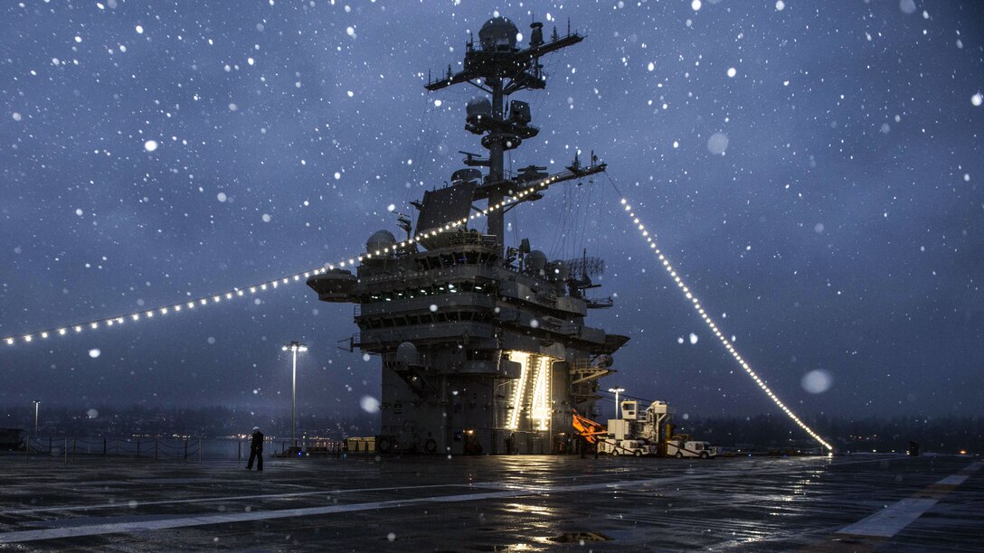 Snowflakes, prominent against a dark blue sky, fall on a ship's flight deck.