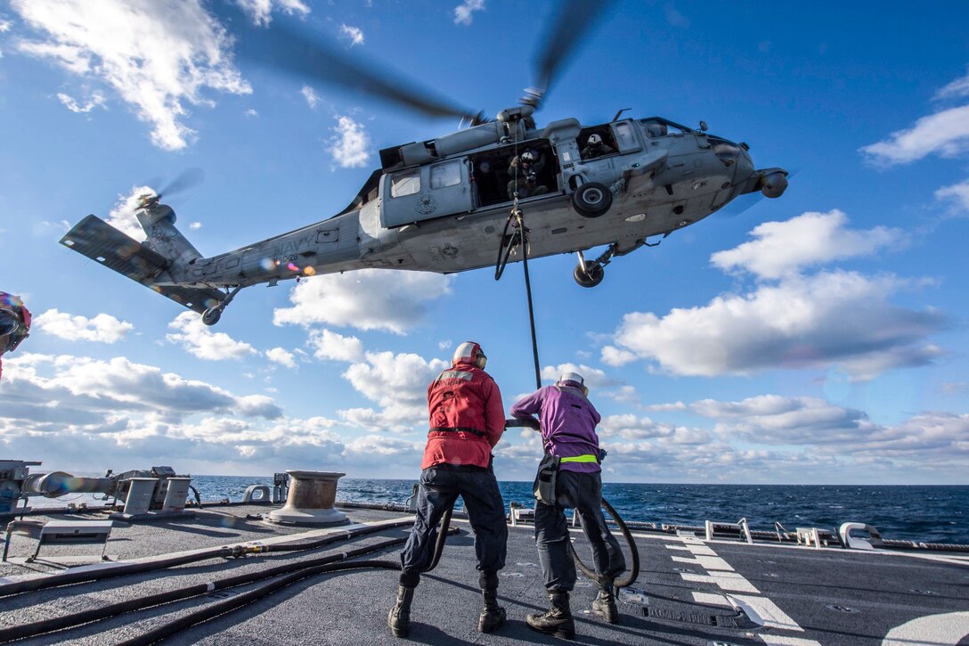 Two sailors on a ship deck feed a fuel line to a helicopter hovering overhead.