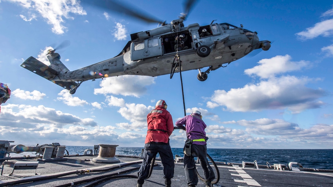 Two sailors on a ship deck feed a fuel line to a helicopter hovering overhead.