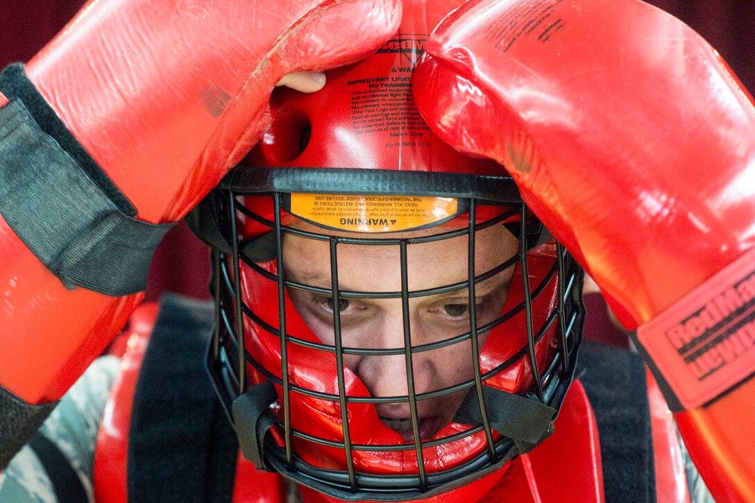 An airman wearing red protective gear adjusts the red helmet with a caged face piece that he's wearing.