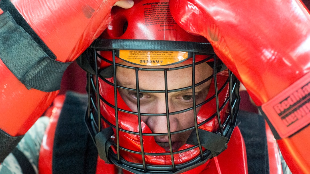 An airman wearing red protective gear adjusts the red helmet with a caged face piece that he's wearing.