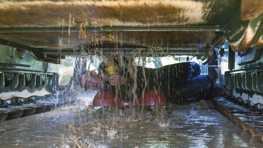 A Marine lies on his side under a vehicle as water pours in front of him.